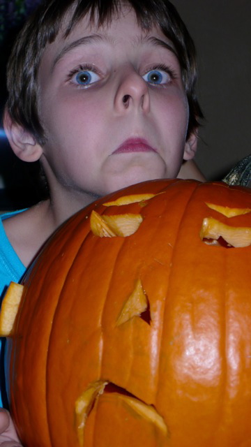 A child makes an exagerate frown. She holds a carved jack-o-lantern with a similar sad face.