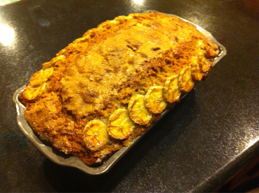 A loaf of banana bread still in the pan. It has a split top, a golden brown color, and two stripes of shingled sliced banana as a decorative top.