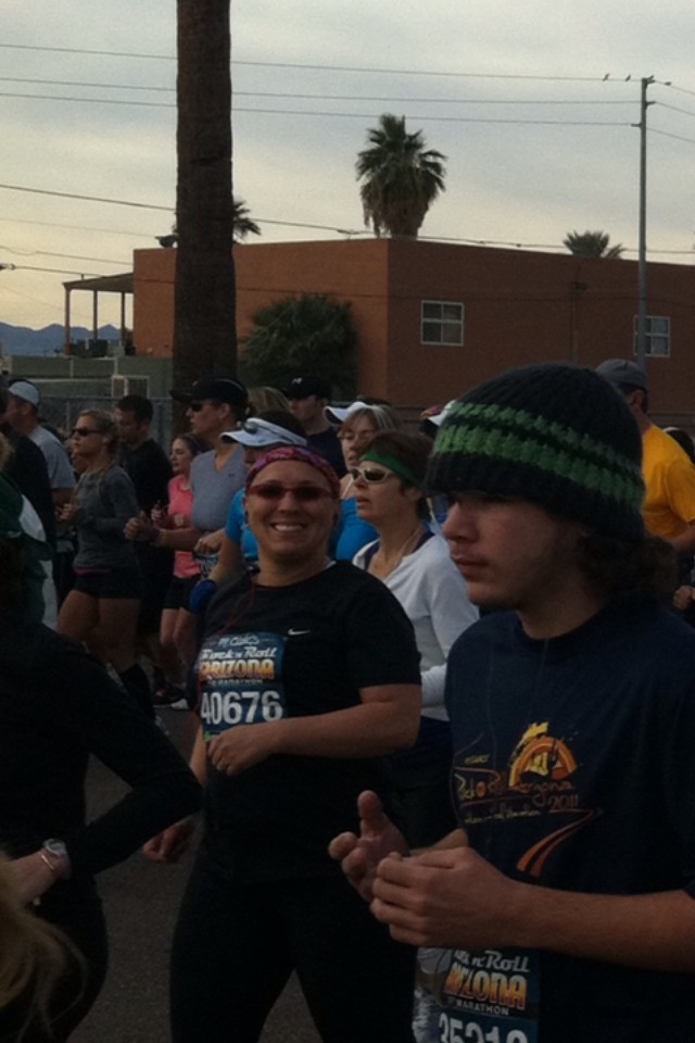 A crowd of people begins to jog past the staring line. They wear Phoenix Rock and Roll Marathon bibs. Mamie is seen smiling at the camera in center frame.