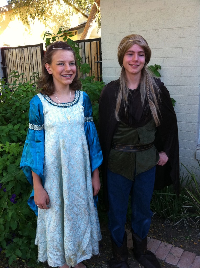 Two teenagers pose in halloween costumes. One wears a blue and silver full length dress and a silver pendant. The other wears a cloak, jerkin, pants and boots, and has a wig with long braids.