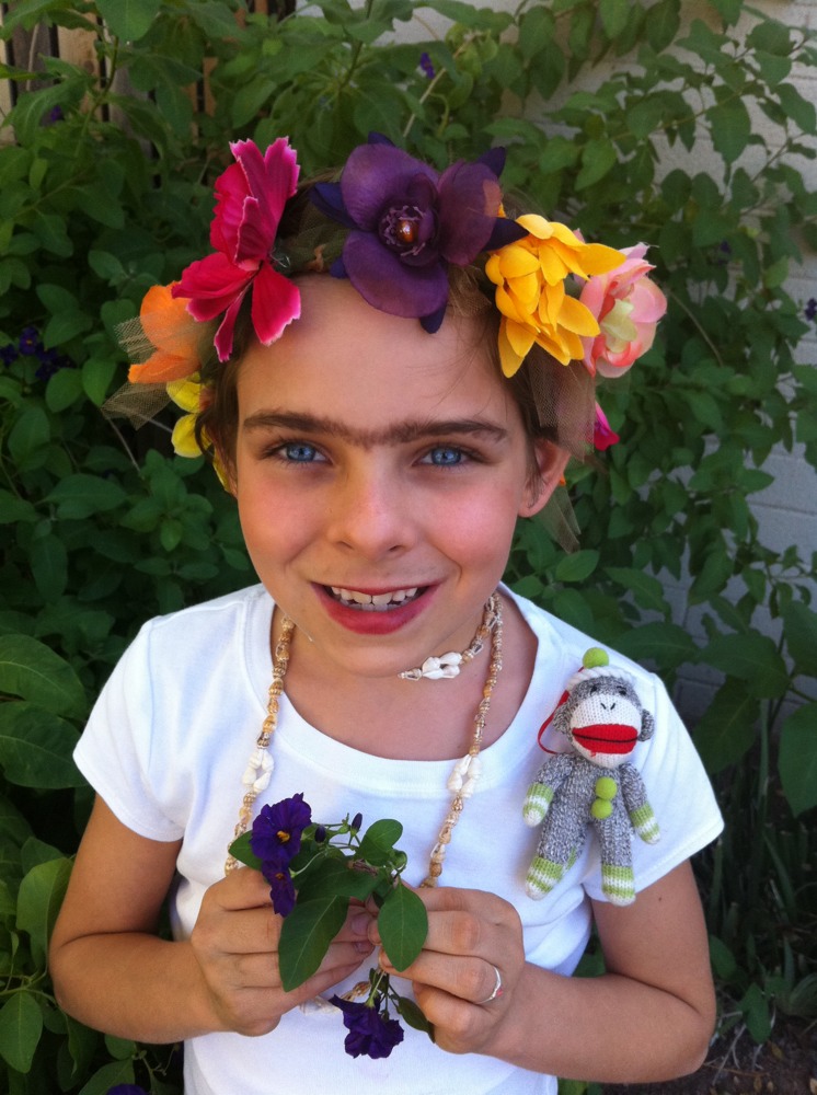 A young girl smiles for the camera. Her head is crowned in a wreath of bright flowers. She has a unibrow and a small sock monkey pinned to her shoulder.