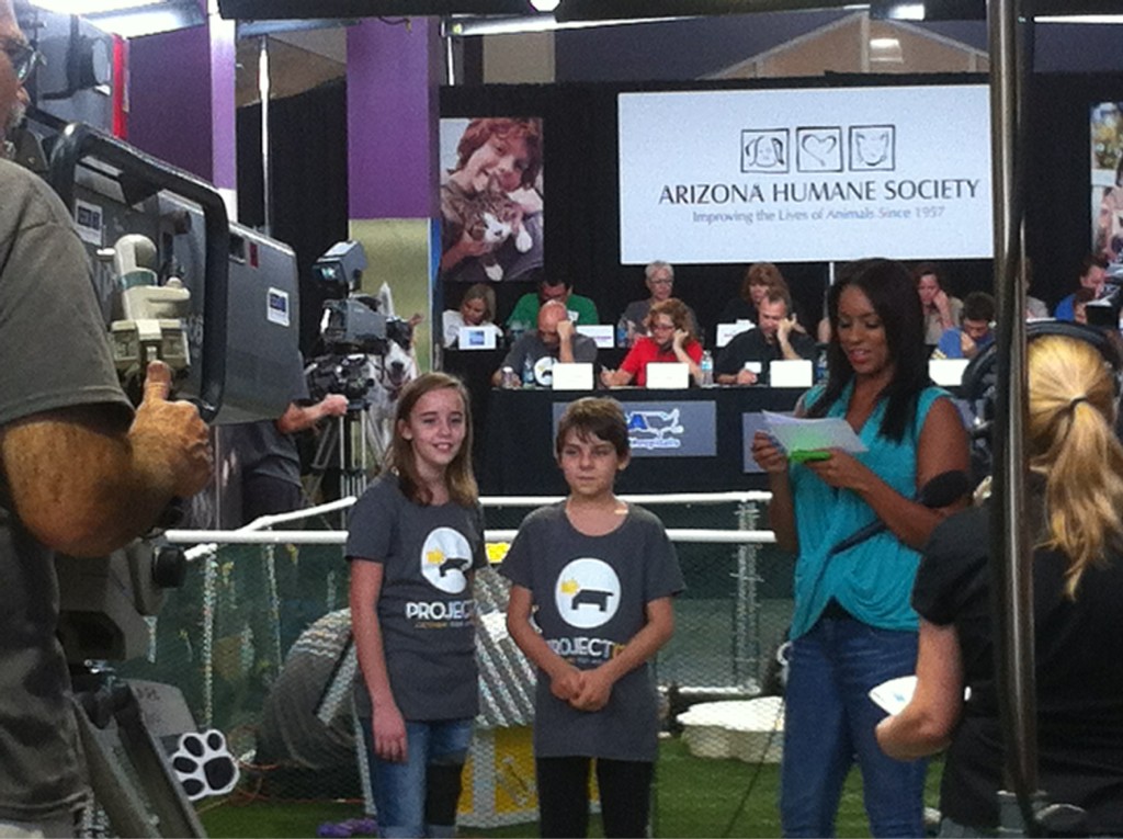 Two children stand in front of a television camera while the news caster reviews her notes near by. They are in a big gymnasium full of people answering phones.