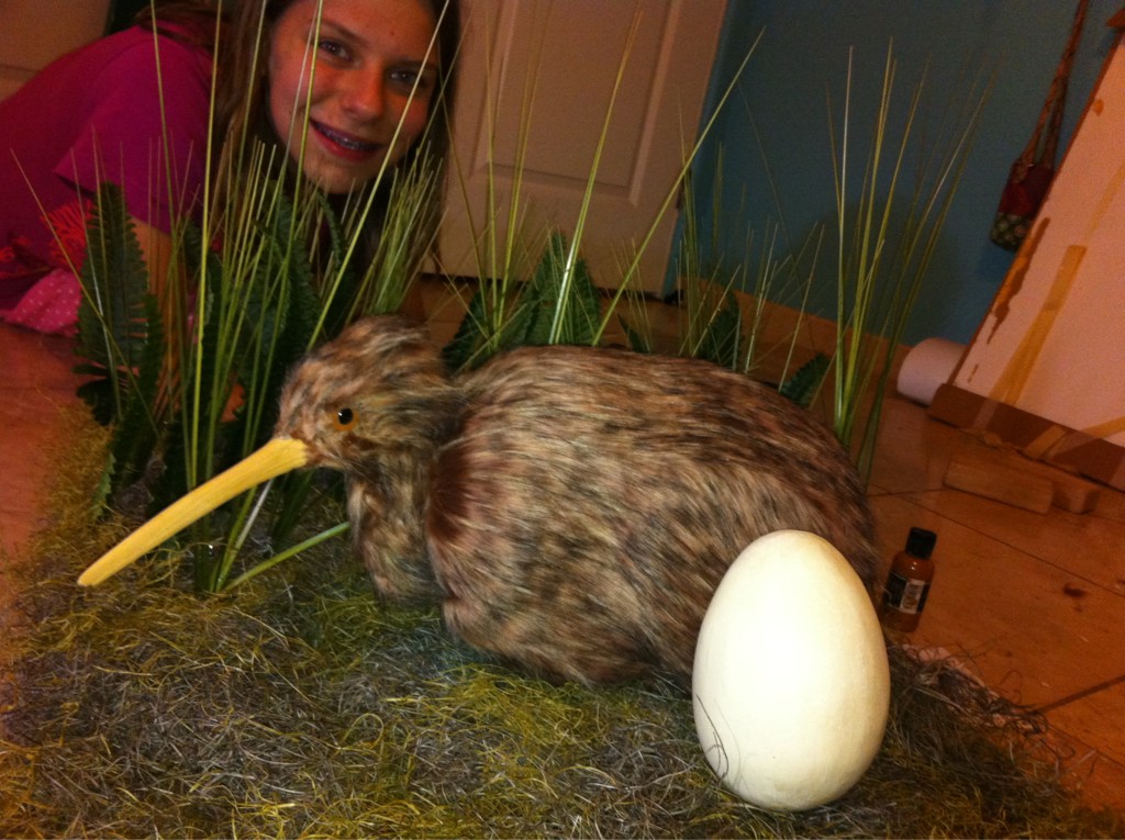 A teenager smiles behind a diorama of a Kiwi bird and egg. The bird is very lifelike with fine hair-like feathers, a long slim beak, and glassy black eyes.