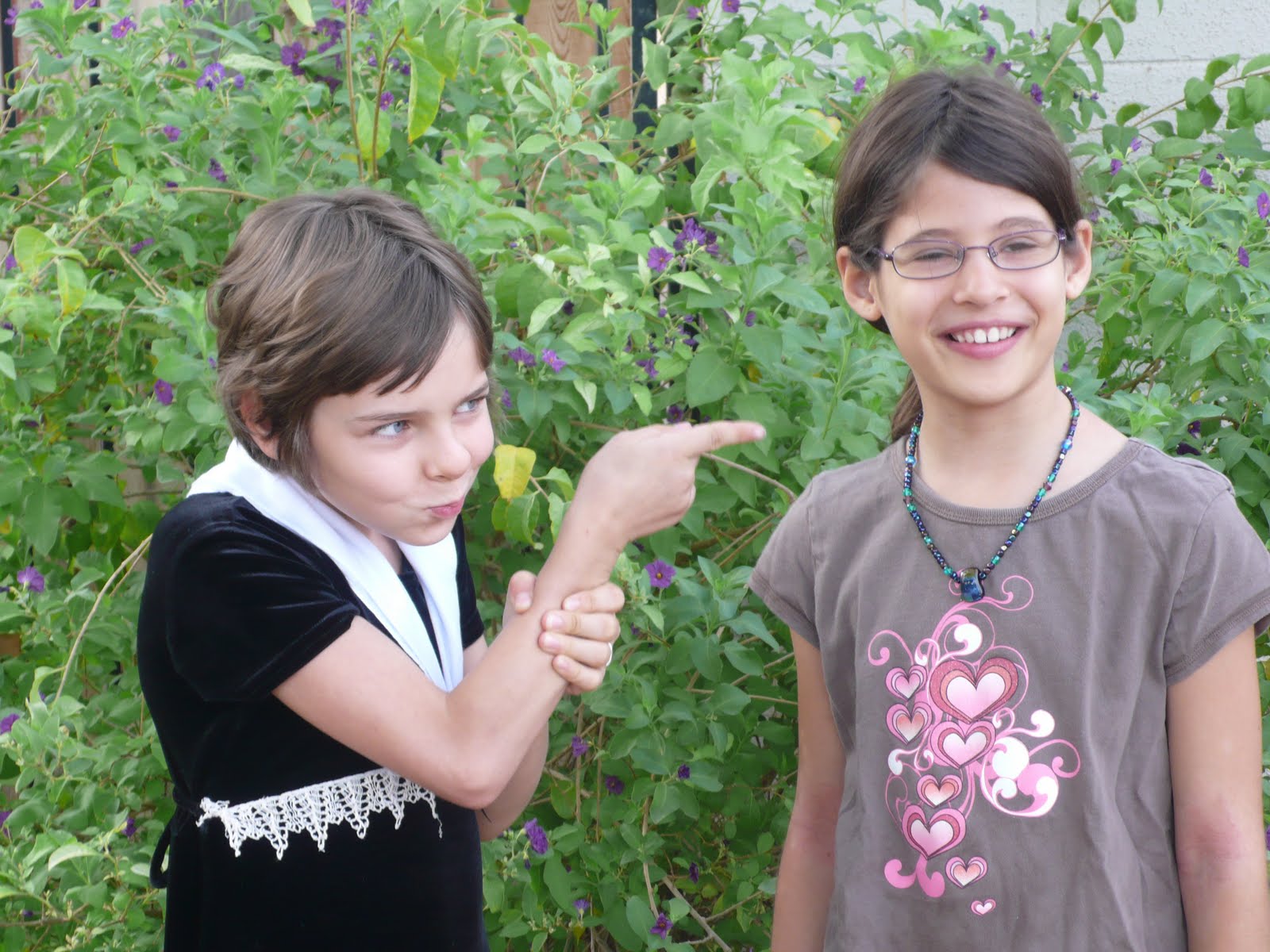 Two girls stand in front of greenery. One is smiling nicely. The other is pointing at the first with a funny/suspicious look on her face.