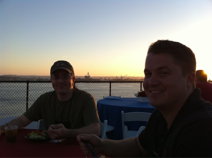 Two men sit at a table eating dinner. The sun is setting over the water behind them.