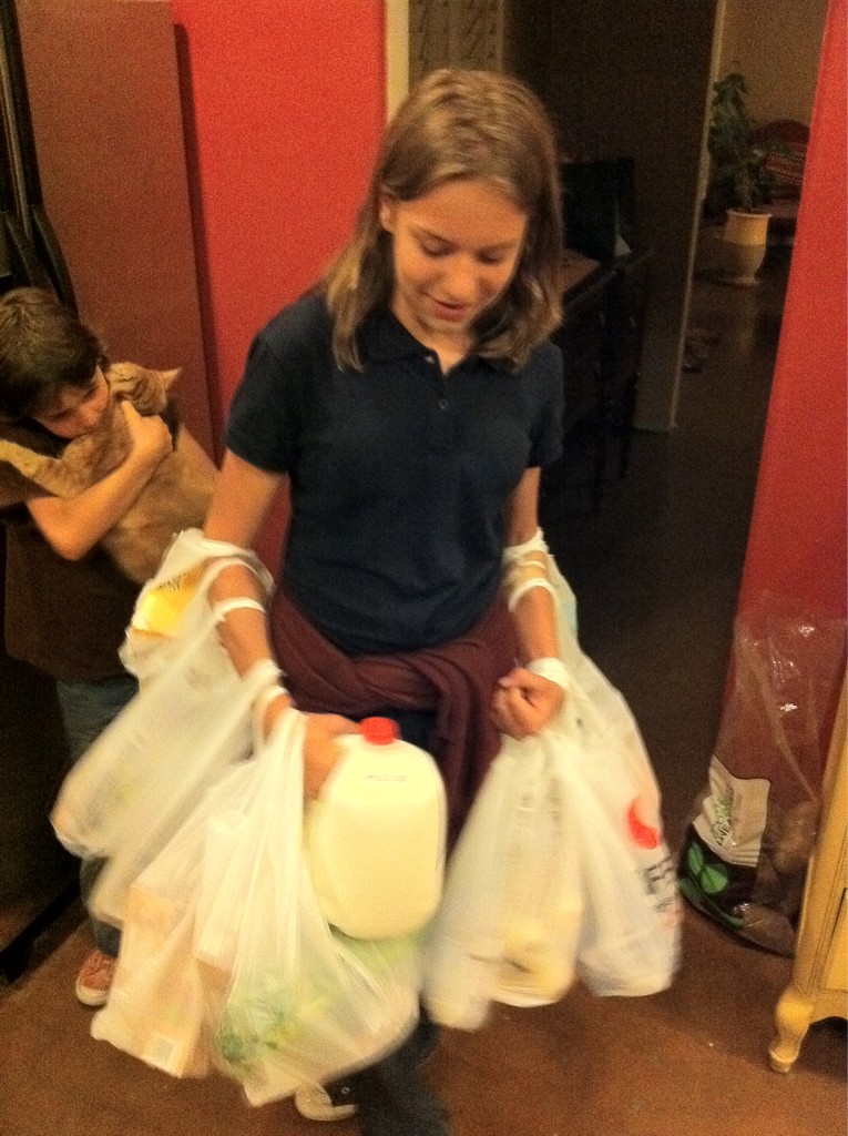 A teenager dressed in their school uniform carries a gallon of milk and perhaps a dozen plastic grocery bags all at once. The bags are hanging from their arms.