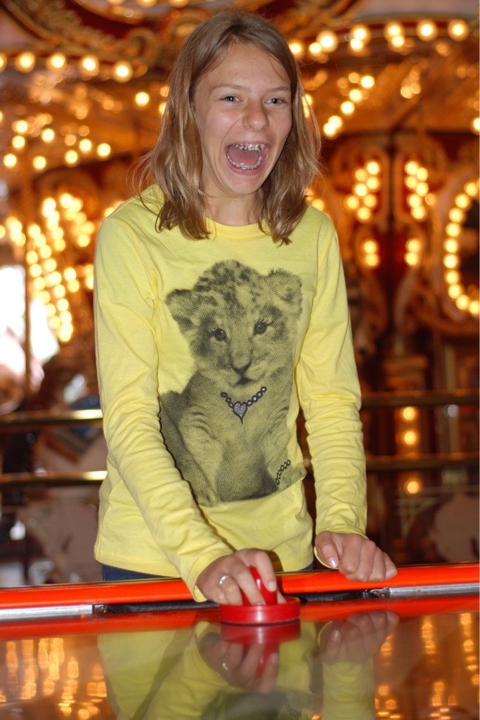 A teenager with a huge open-mouthed smile of excitement. They stand at the end of an air hockey table with bright carnival lights behind them.