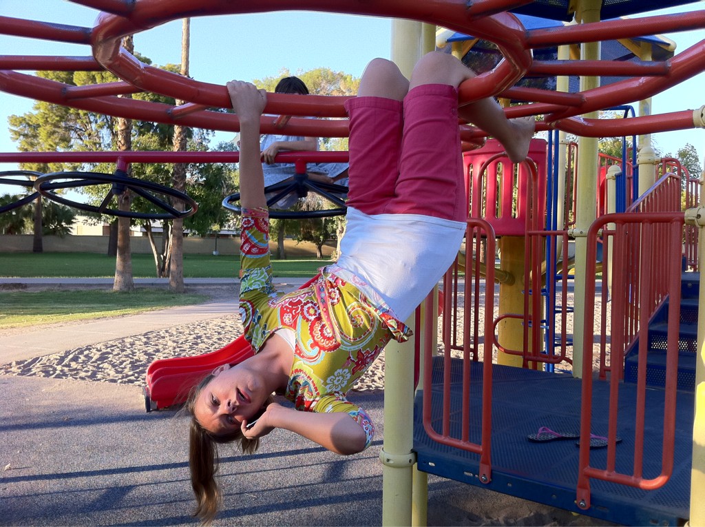 A teenager hangs upside down from a playground structure while holding a mobile phone to their ear.