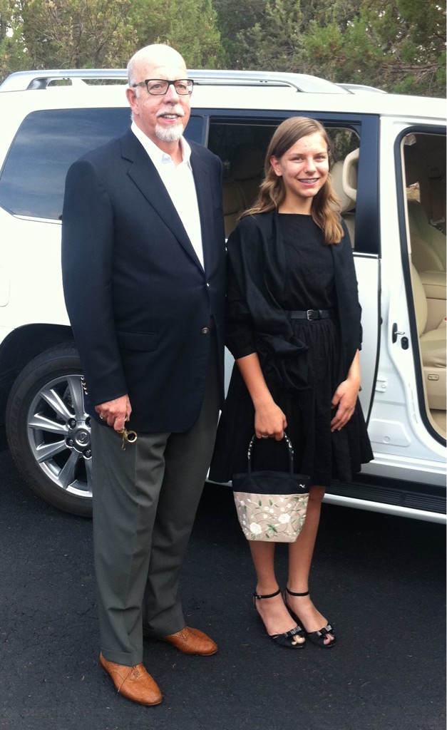 A teenager in a black dress with a pretty silk flower-patterend purse stands beside an older man in a suit. They are about to enter the open door of a car.