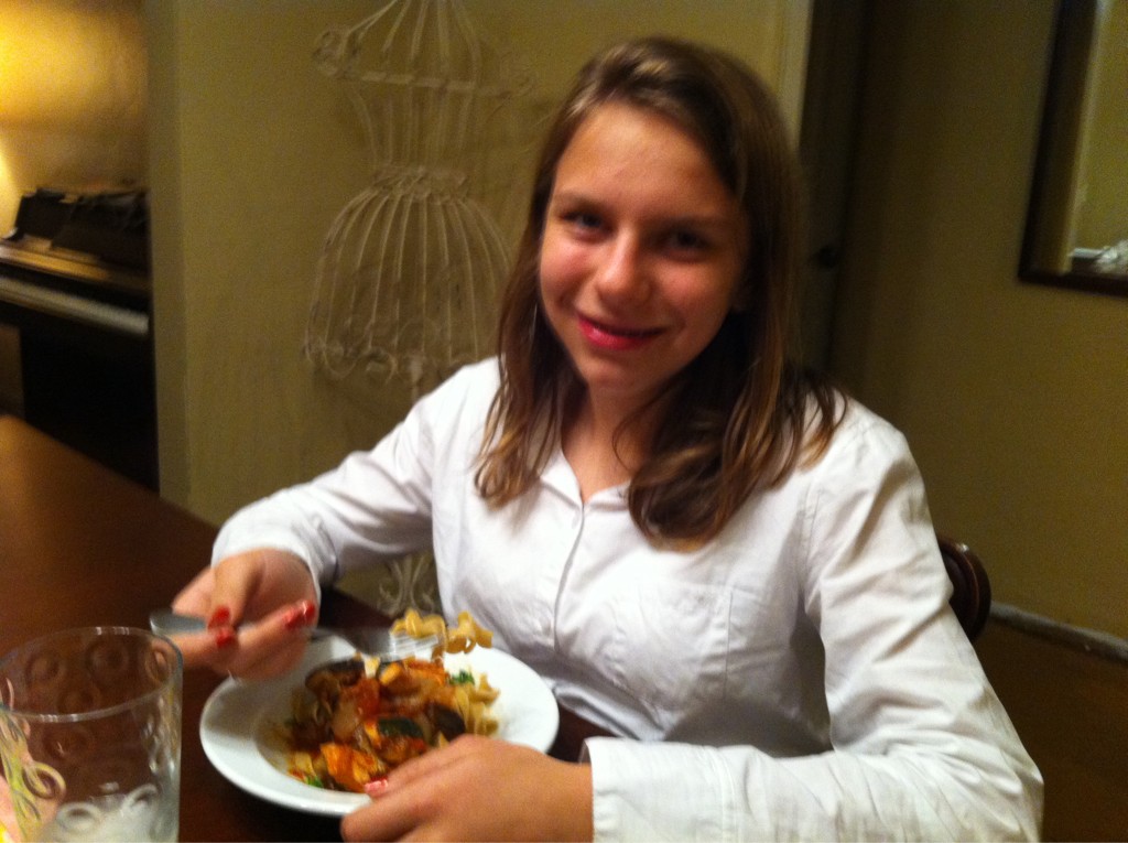 A teenager sits at a dining table smiling for the camera. They are wearing a white button-up collared shirt and have long straight dirty-blonde hair. There is a large bowl of rustic ratatouille on the table.