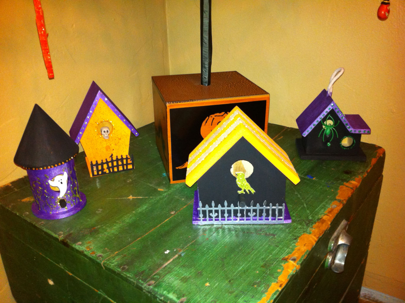 A distressed green chest with four small birdhouses arranged haphazardly on top. Each house is a different structural design, and each is painted in haloween colors.