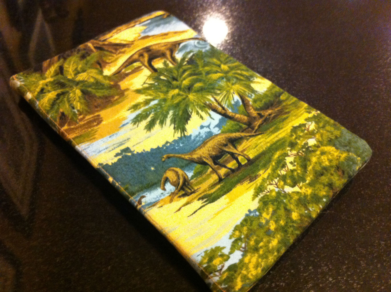 A Kindle laying on a black countertop. The Kindle is covered in a cloth case with a dinosaur and primitive vegetation print.