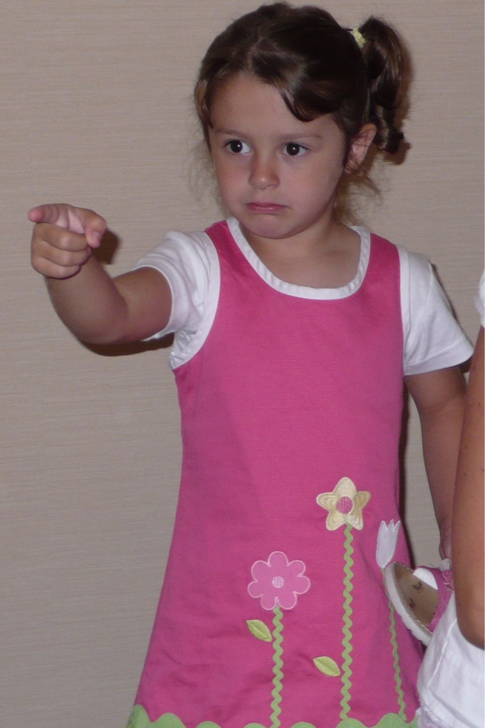 A little child in a pink dress points at someone off camera with a stern look on their face.