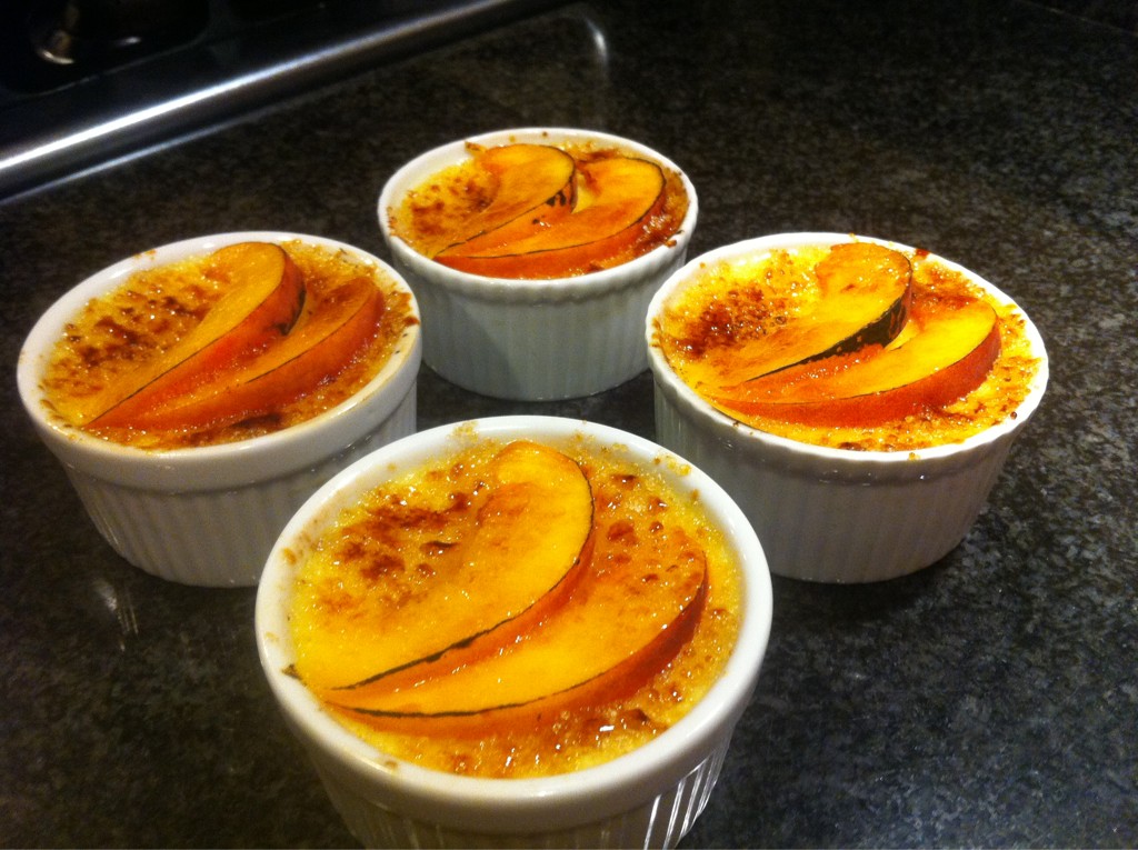 Four white ramekins of Crème Brûlée sit on a black countertop. Each has two slices of fresh peach decorating the top.