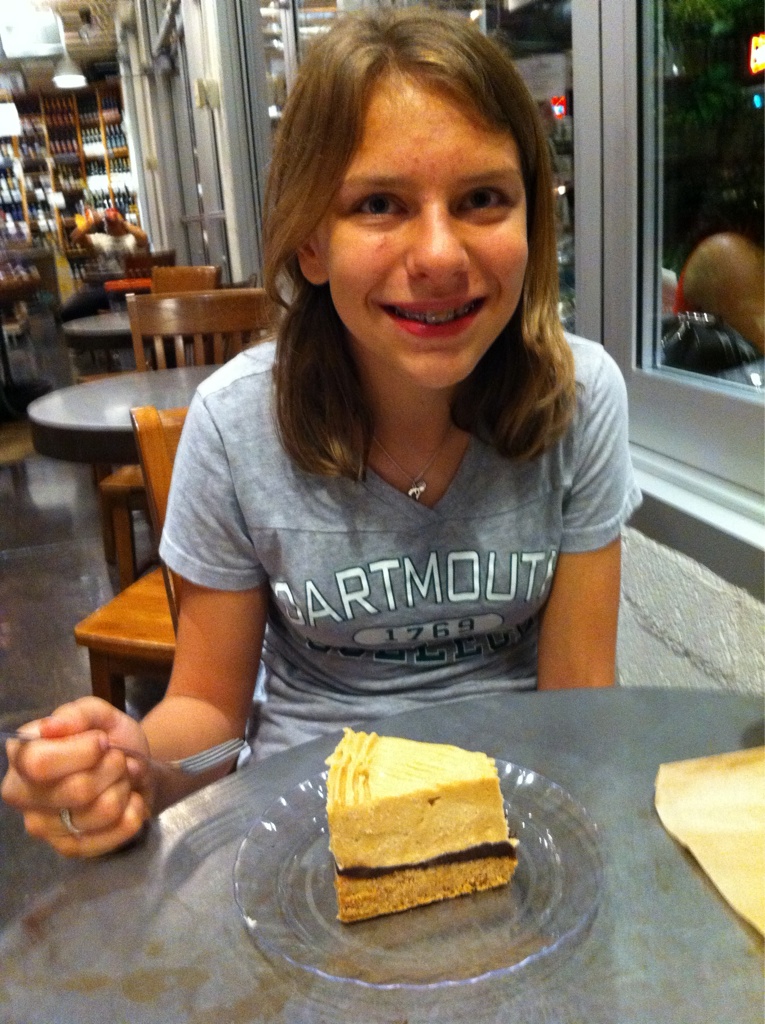 A teenager wearing a Dartmout t-shirt sits at a reastaurant table smiling for the camera. They hold a fork. Before them on the table is a large slice of peanut butter pie.