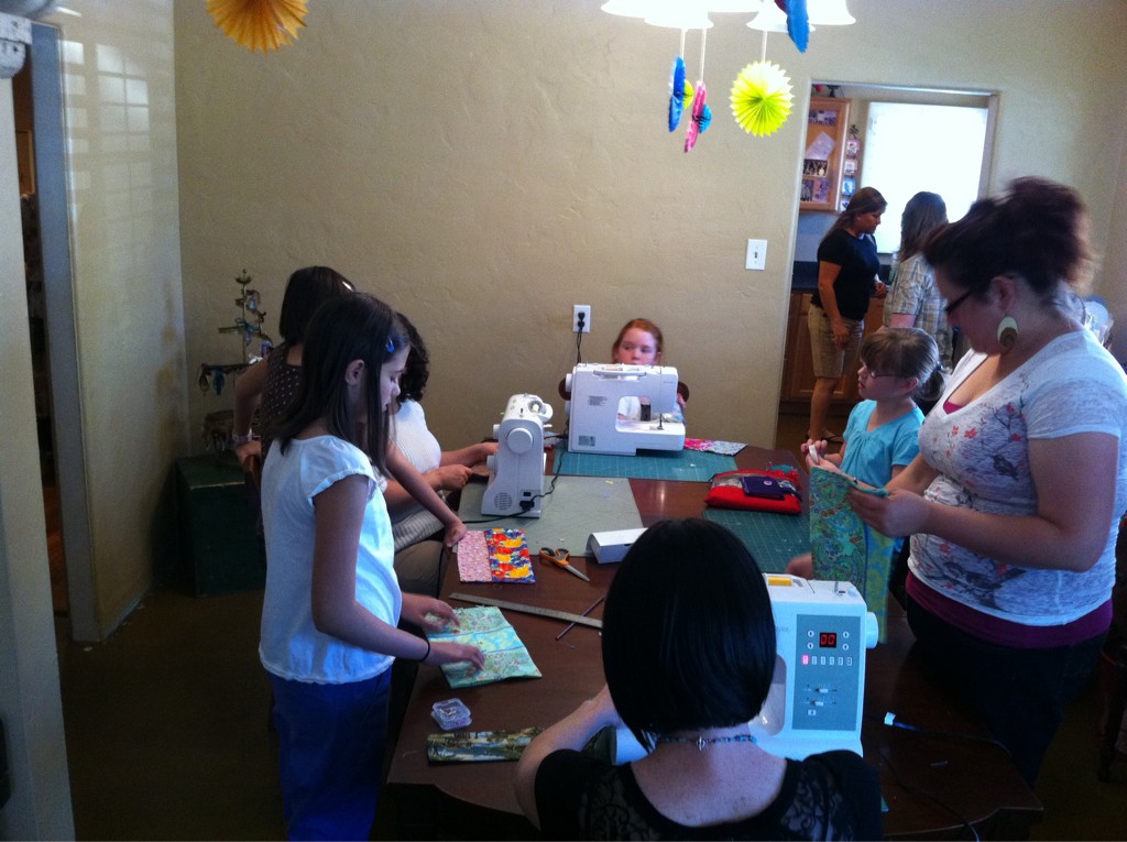 A room full of middle-school aged children and a few adults around a large table. There are several sewing machines on the table.