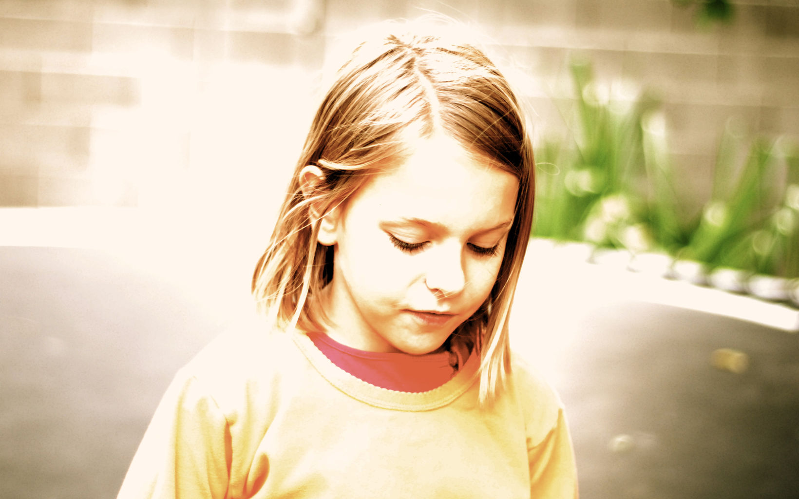 An over-exposed photo of a child with medium length hair wearing a yellow shirt over an orange shirt. They are looking down with interest at something off camera.