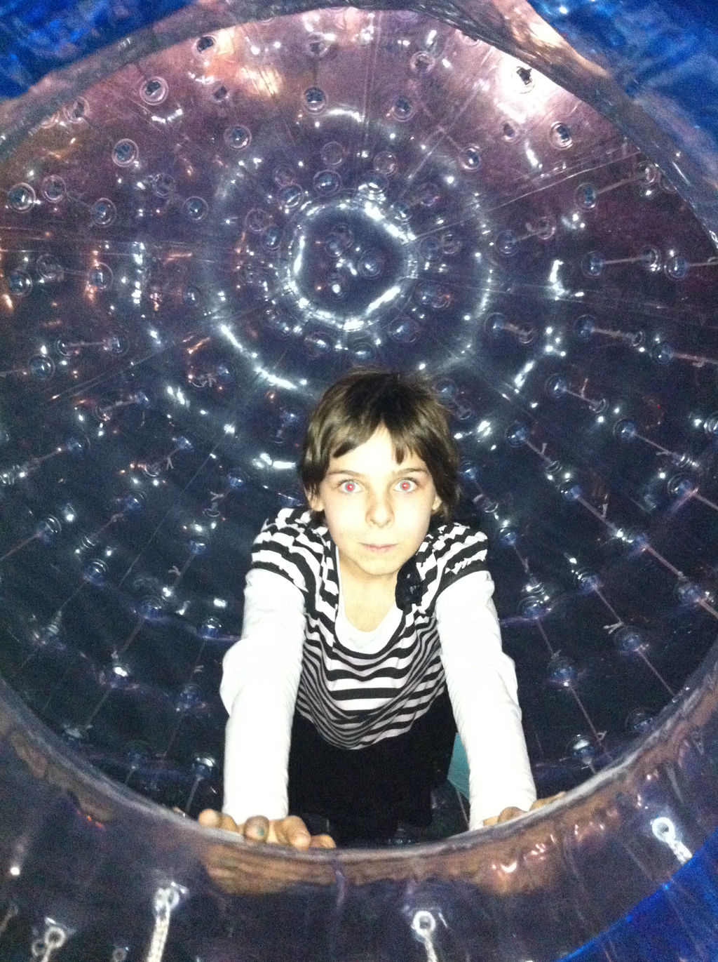 A young girl looking straight at the camera through the opening in an enormous human-sized inflatable "hamster ball".