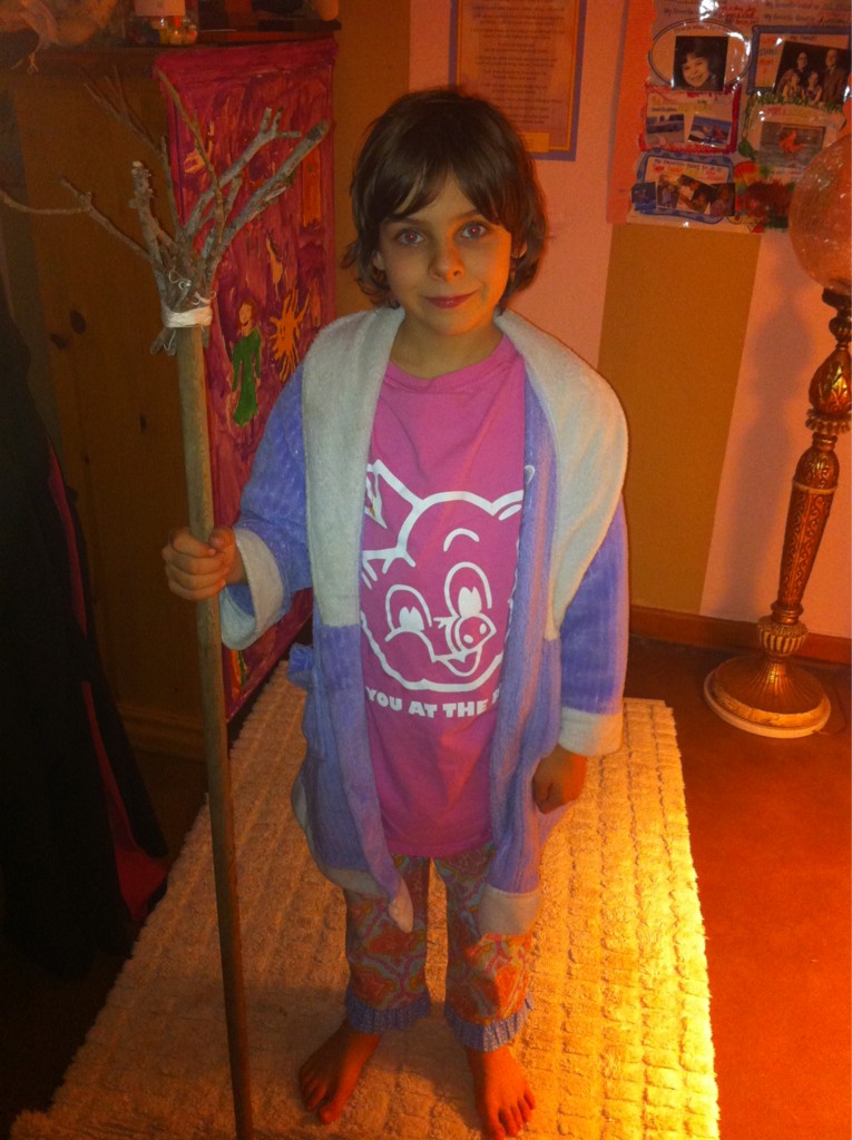 A young girl wearing a bath robe and holding a broom handle with twiges collected from the back yard tied in a bundle at the end.