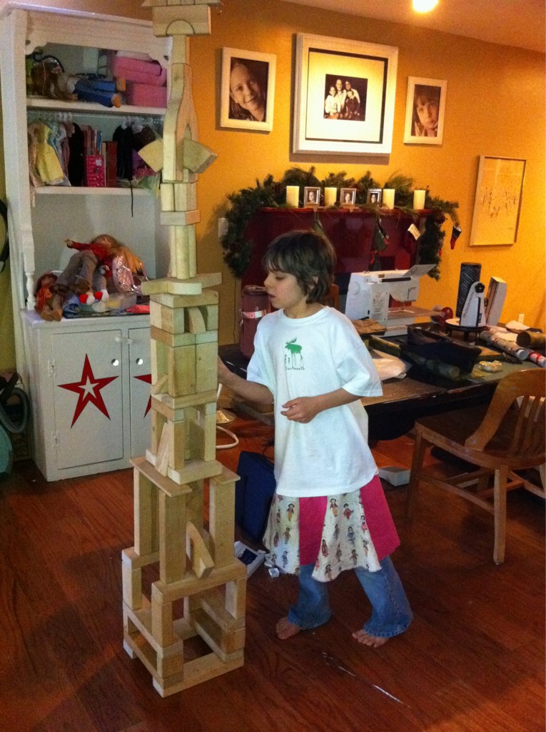 A young girl puts the finishing touches on a tall tower made of wooden toy blocks. The tower reaches the ceiling.