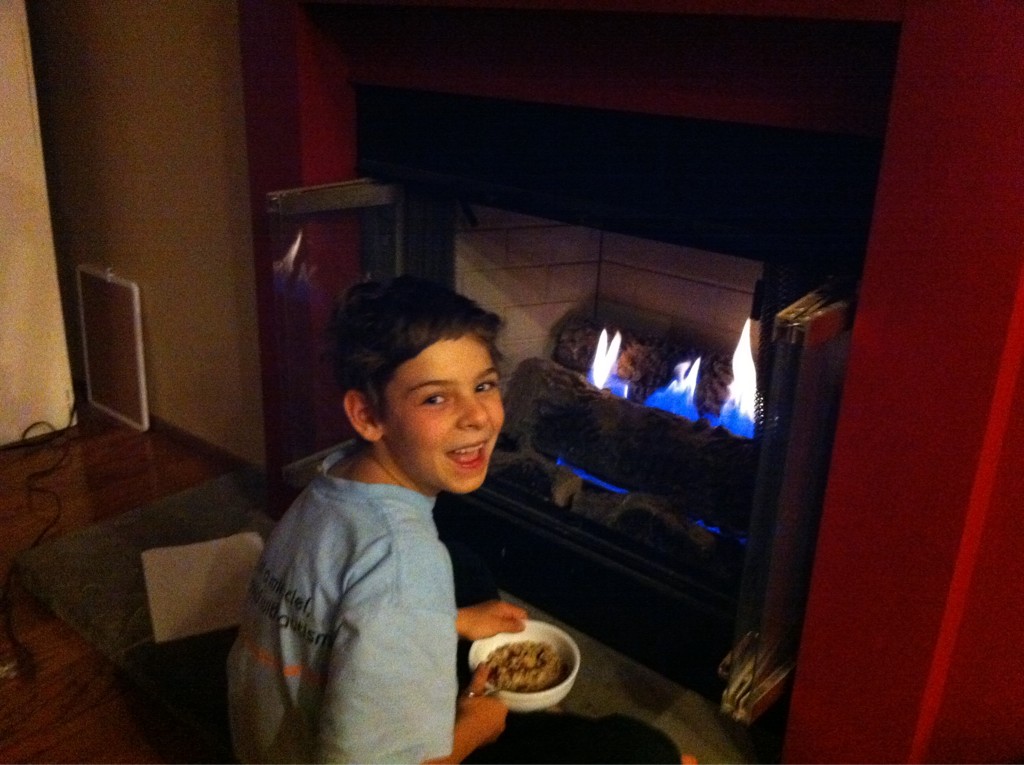 A young girl sits very close to the fire in the fireplace holding a bowl of hot oatmeal. She smiles widely.