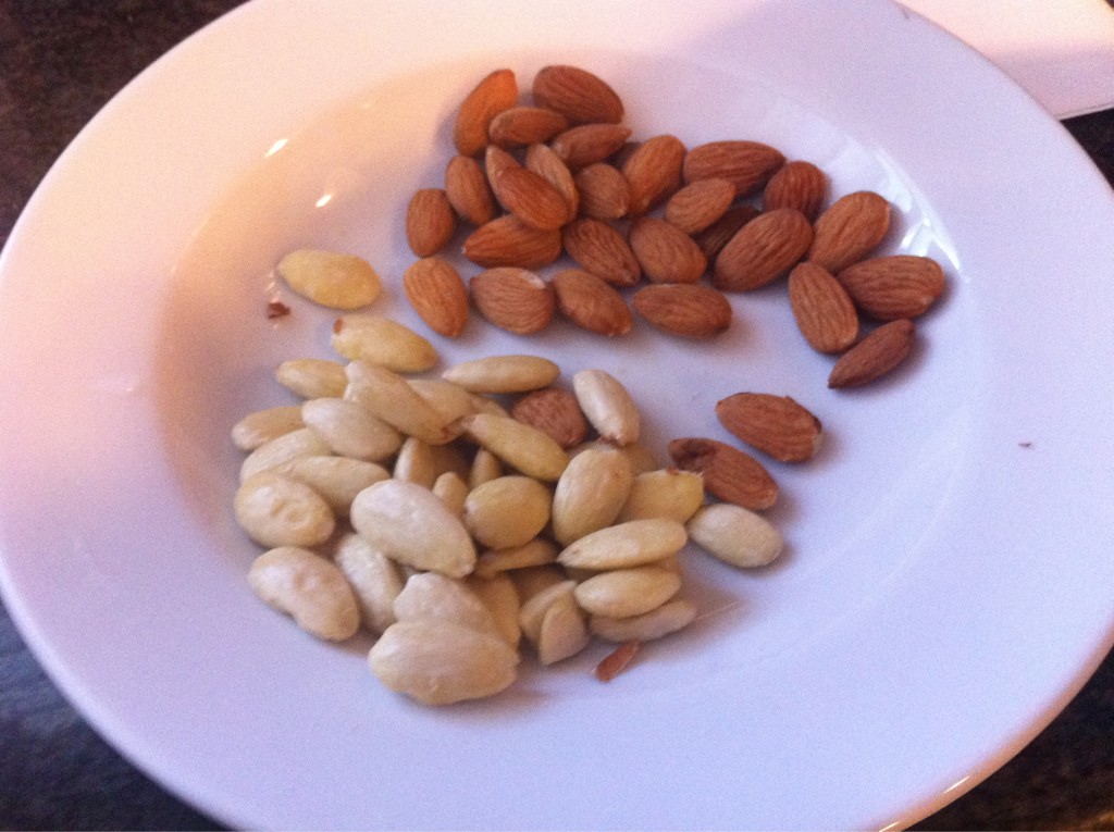 A white ceramic plate has two piles of almonds. In one pile the almonds are glossy and light in color because they have been "peeled"