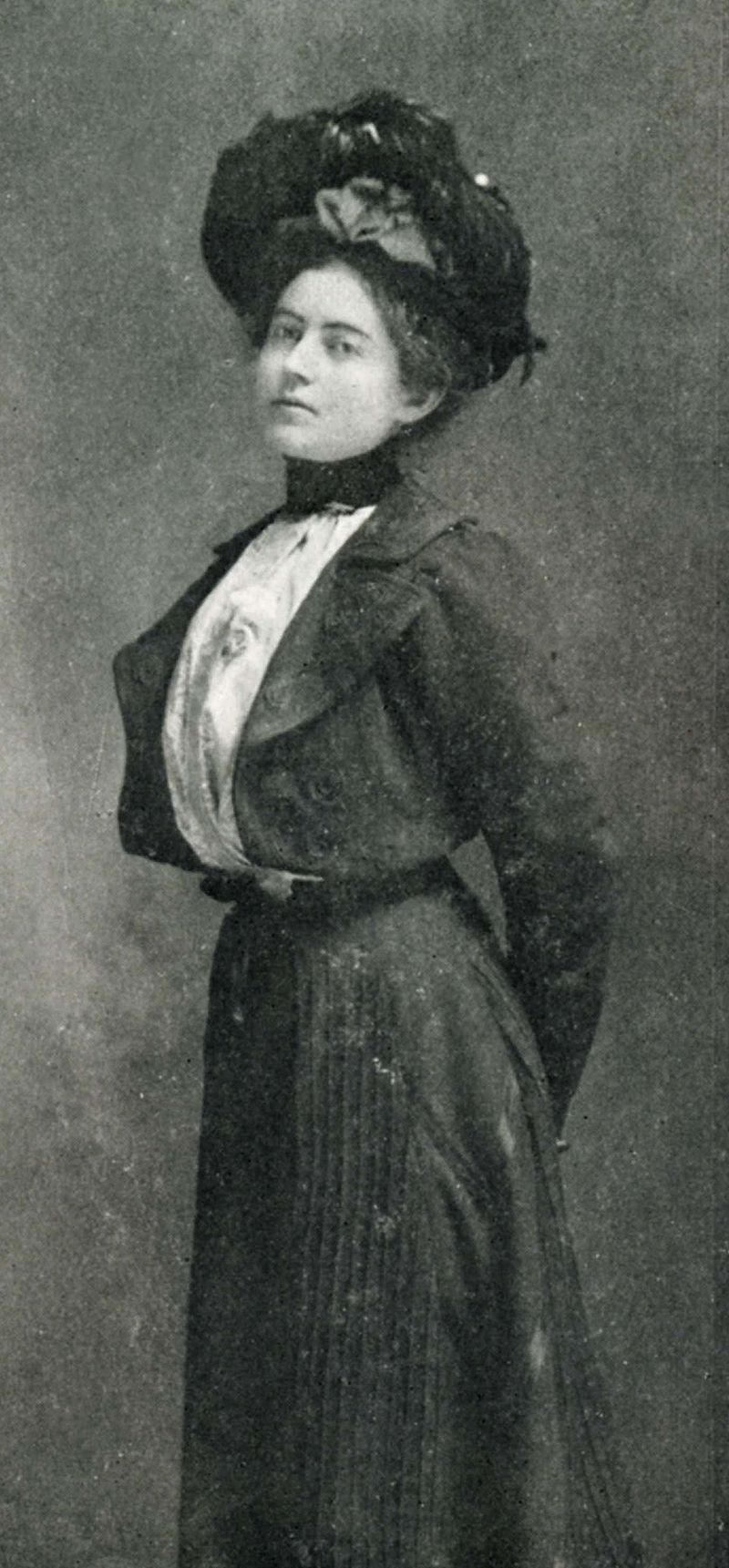 An aging black and white photograph of a young Mary MacLane. She wears 19th century clothing including a large hat, and long full dress. Her arms are behind her back, body erect, head slightly back. She has a defiant look on her face.